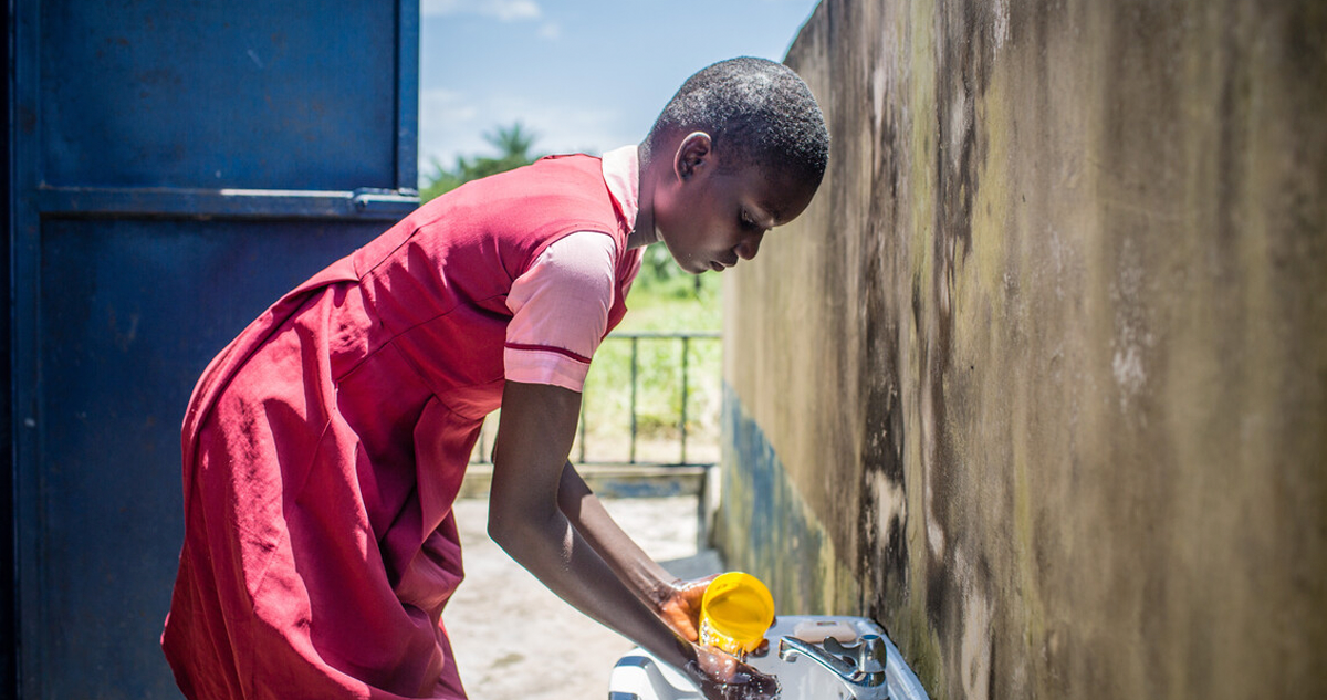 Maria using a cup of water at a sink outside to wash her hands.
