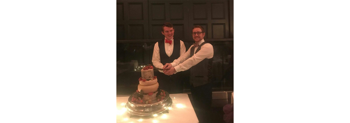 Wil Organ smiling at his husband as they cut their wedding cake together.